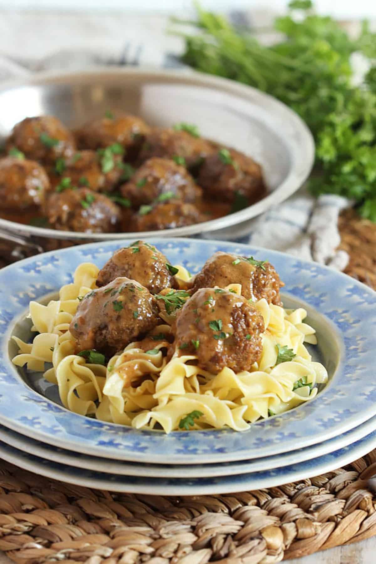 Swedish Meatballs on egg noodles plated on a blue plate with a skillet full of meatballs in the background.