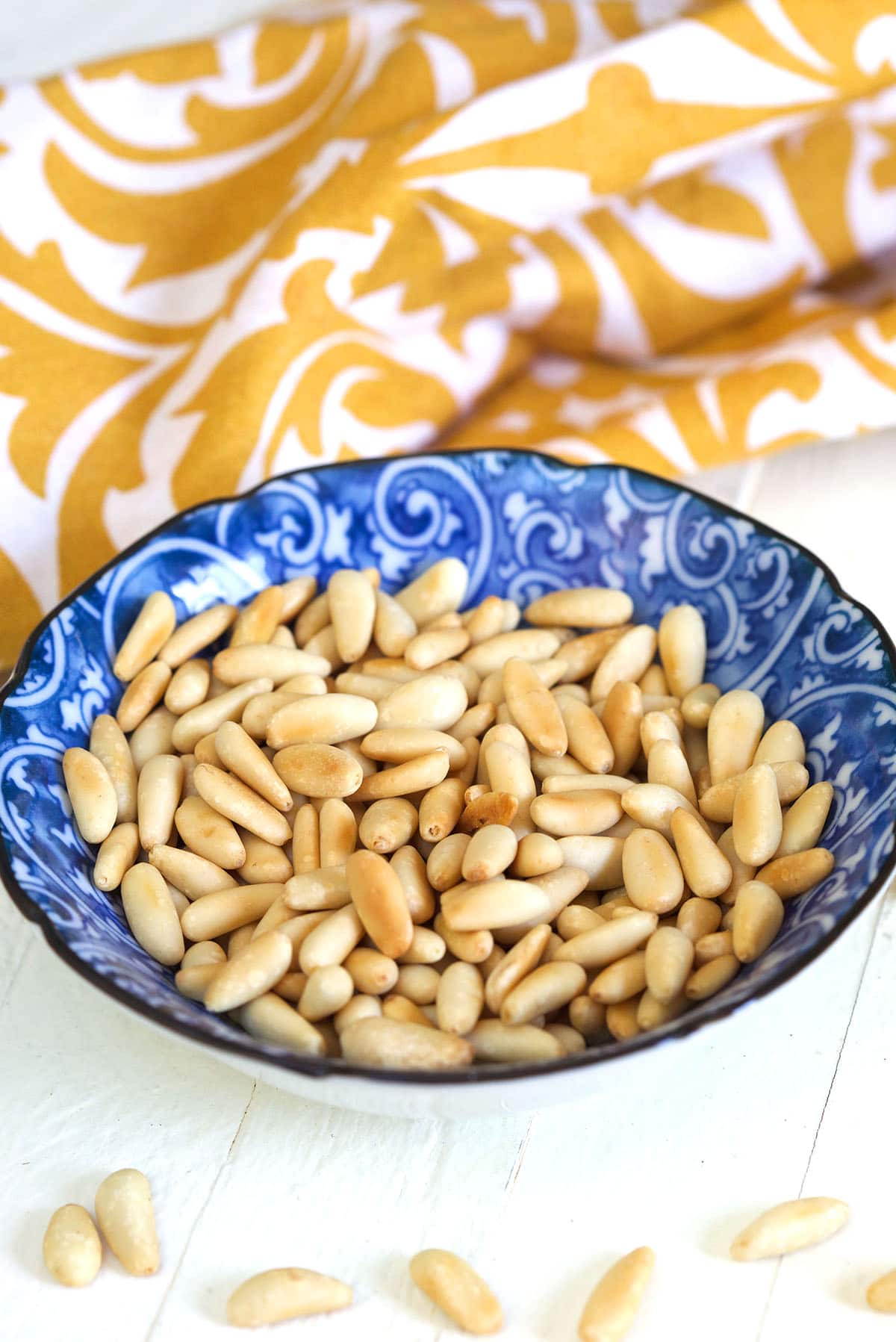 Toasted pine nuts in a blue bowl are placed next to an orange and white cloth. 