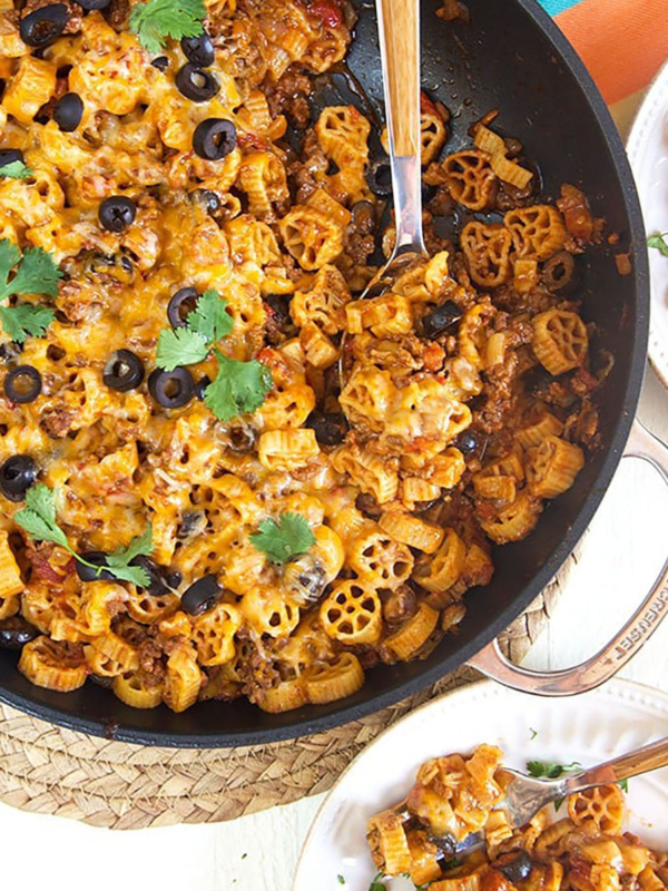 Overhead shot of taco pasta in a black skillet with a wooden spoon on a white background.