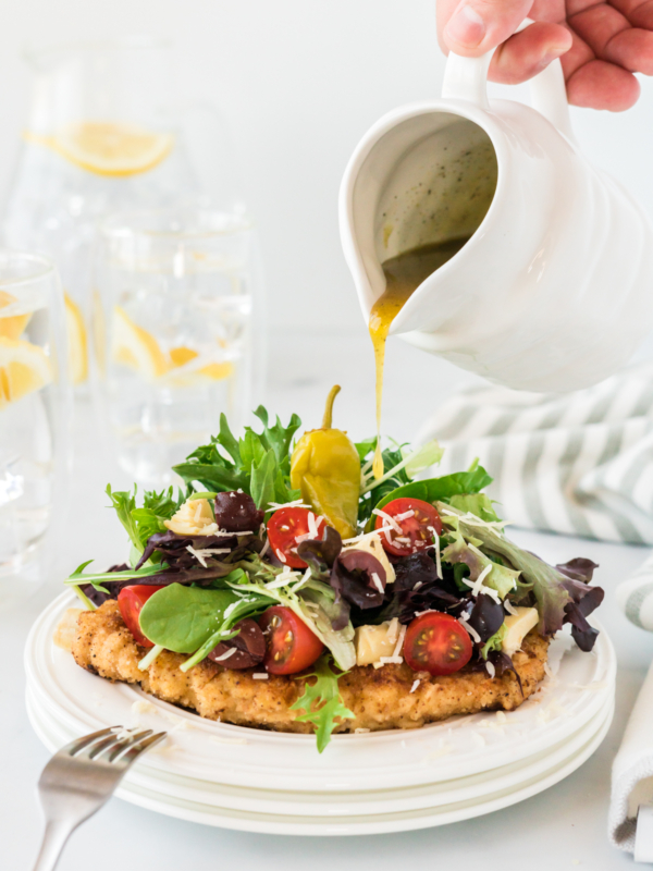 dressing being poured over Chicken Milanese on a white plate.