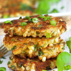Three corn fritters are stacked on a white plate with a sprig of basil.