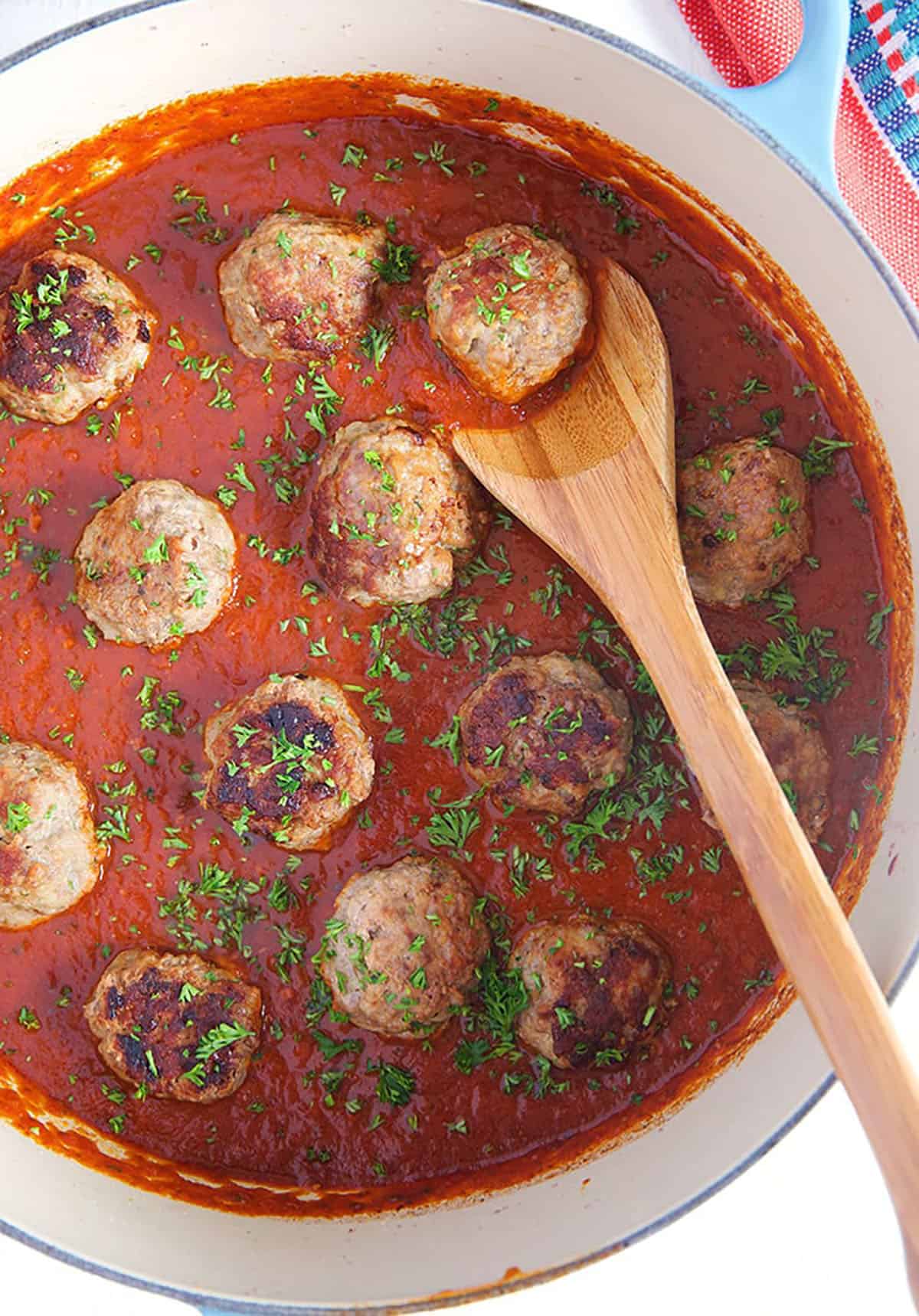 Overhead shot of pan fried meatballs in a dutch oven with a wooden spoon and tomato sauce.