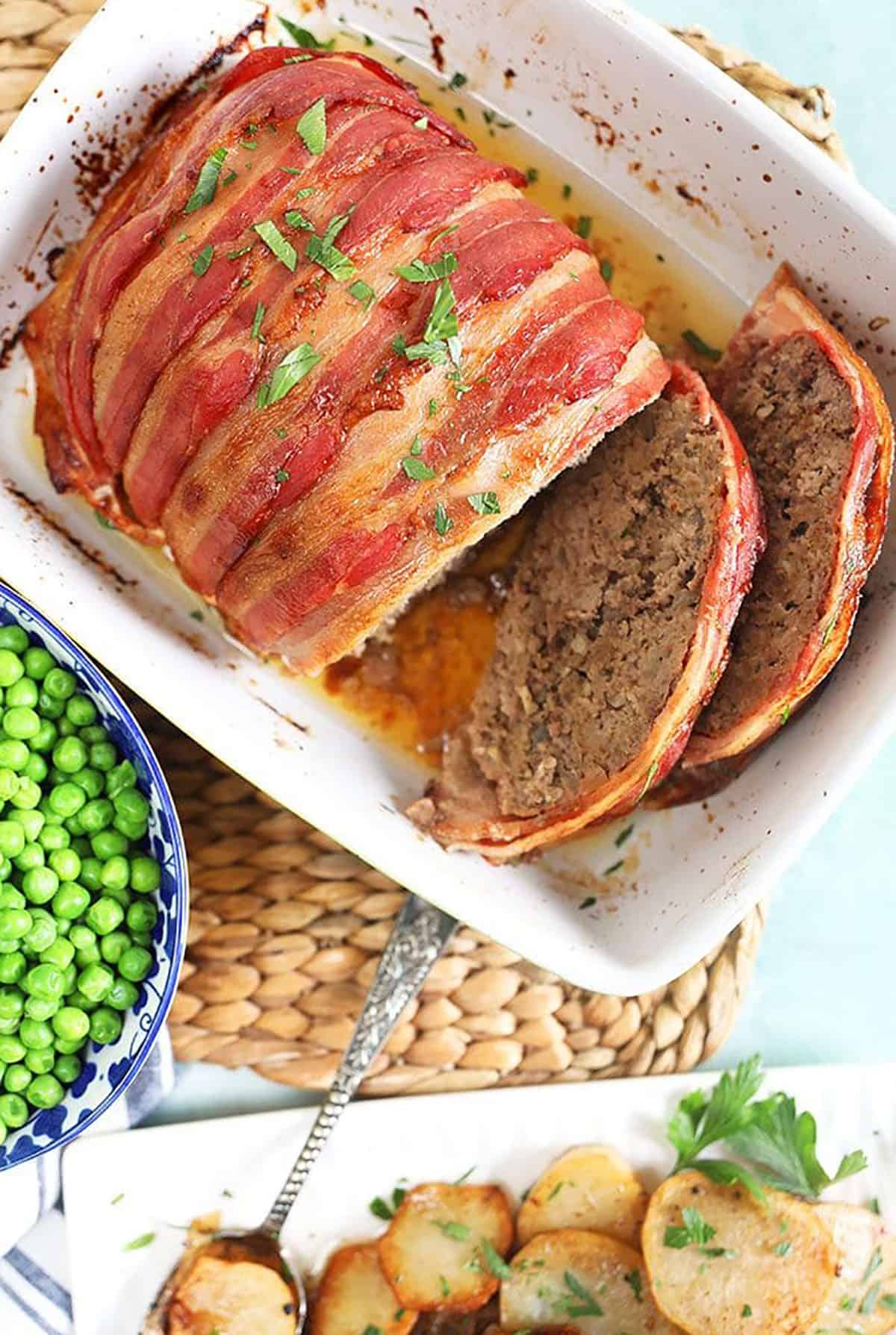 BAcon wrapped meatloaf in a baking dish sliced with a plate of potatoes.