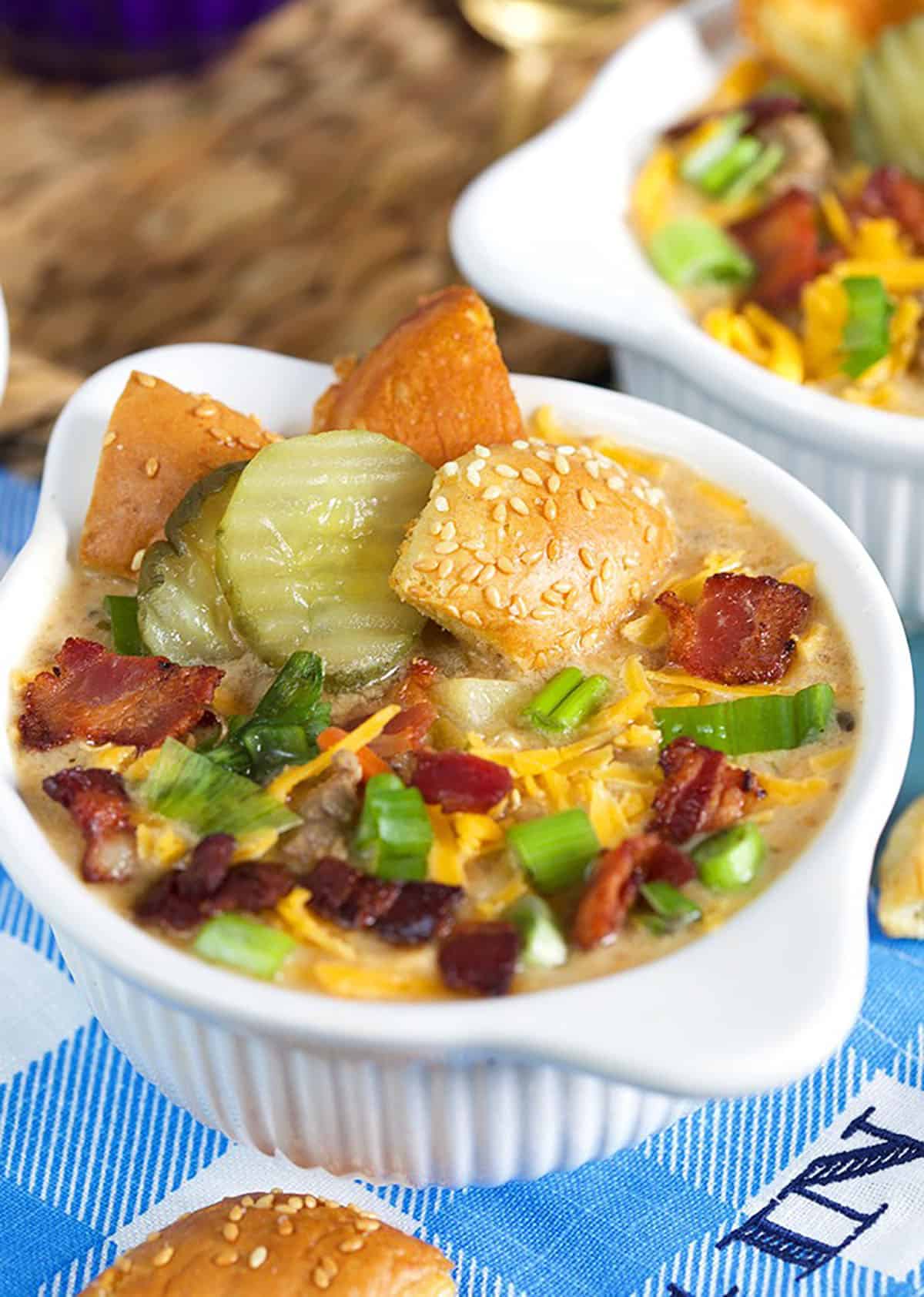 Close up shot of Bacon Cheeseburger Soup in a white bowl with pickles burger bun croutons, bacon and cheese.