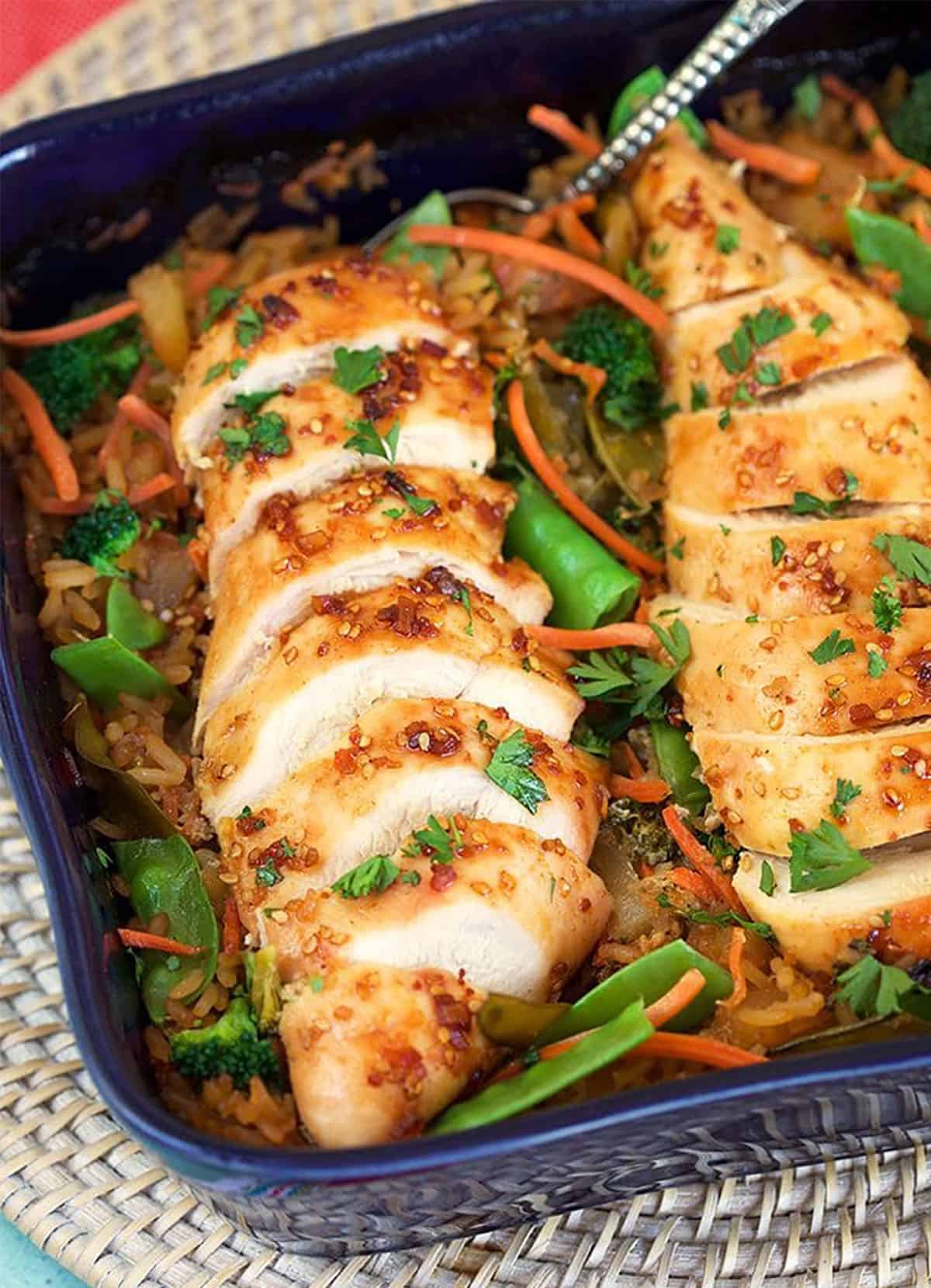 Close up of sliced baked chicken breast in a casserole dish with rice and veggies.