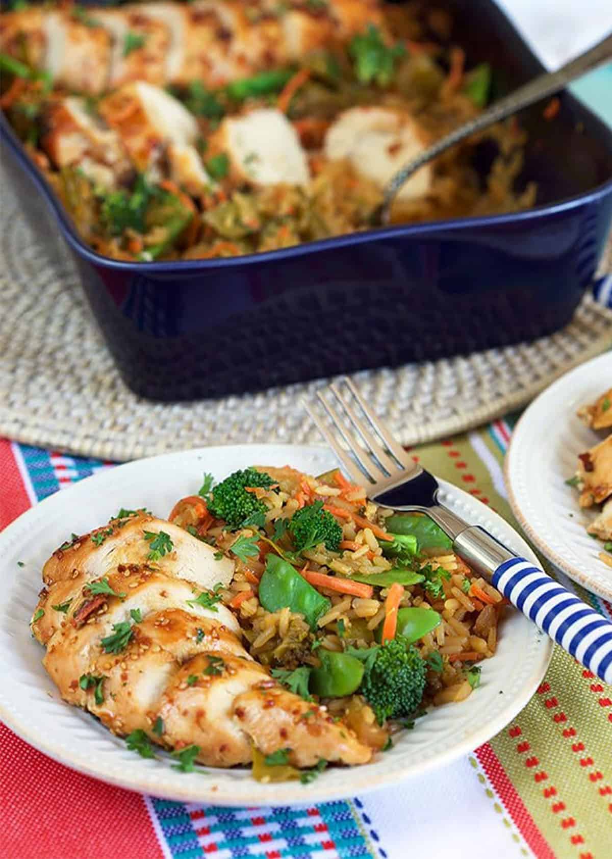 Sliced baked teriyaki chicken on a bed of rice and veggies on a white plate. Teriyaki chicken casserole in a blue casserole behind the plate.