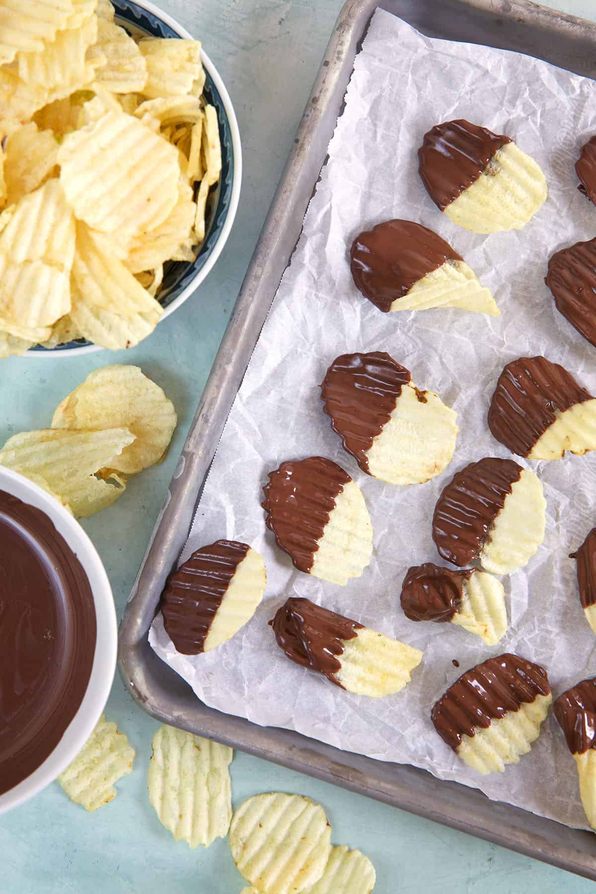 Chocolate dipped potato chips are on a prepared baking sheet, next to more chips and melted chocolate. 