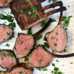 Several slices of grilled lamb are presented on a white surface next to the whole rack.