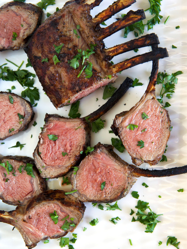 Several slices of grilled lamb are presented on a white surface next to the whole rack.
