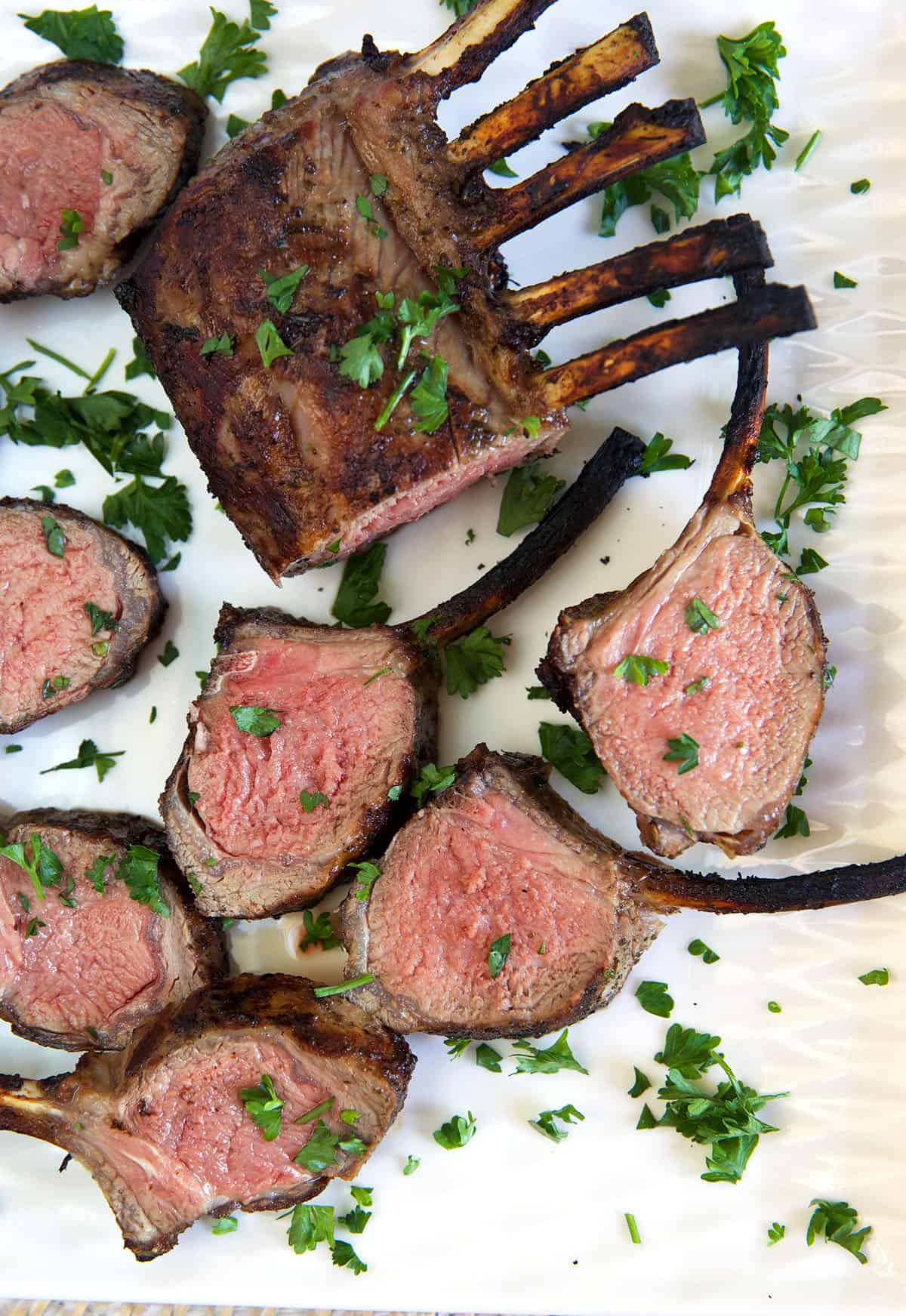 Several slices of grilled lamb are presented on a white surface next to the whole rack.
