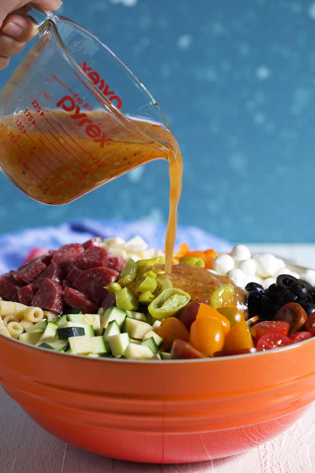 Dressing being poured over Italian pasta salad in an orange bowl.