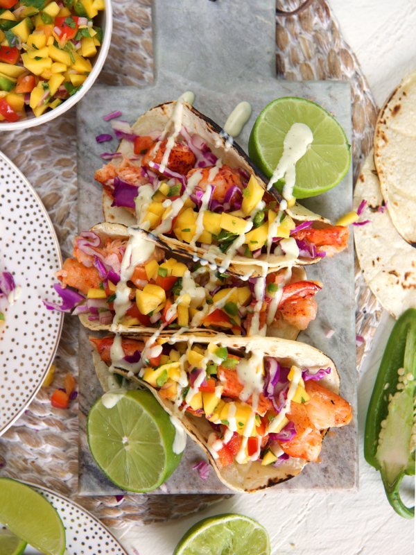 Three lobster tacos are placed on a table next to halved limes.