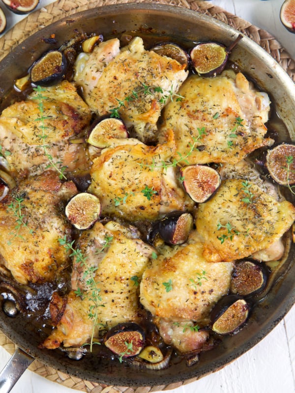 Crispy chicken thighs with roasted figs in a steel skillet on a white background.