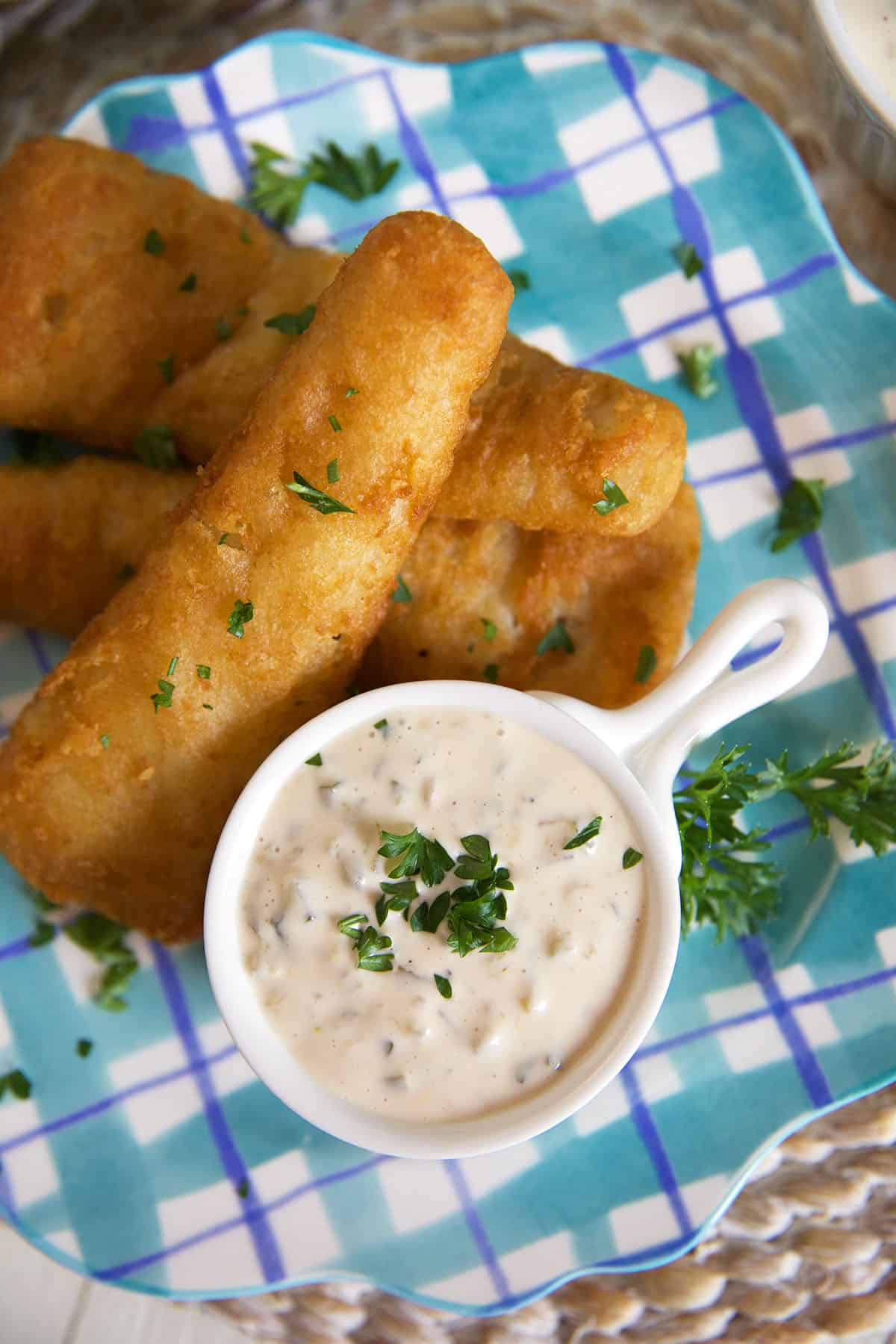 Fish sticks and a white bowl of tartar sauce are on a plaid printed plate. 