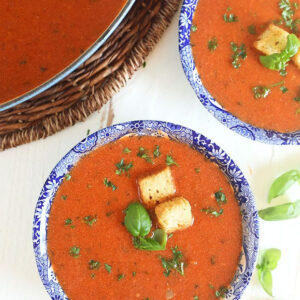 Overhead shot of tomato basil bisque with croutons in a blue and white bowl on a white background.