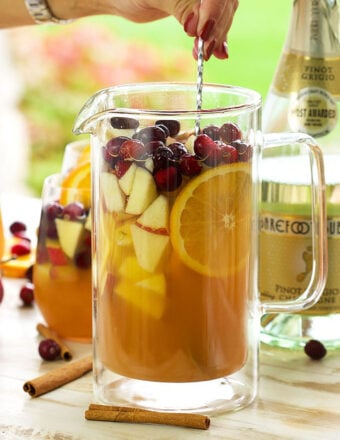 Apple cider sangria in a glass pitcher being stirred with a silver cocktail spoon