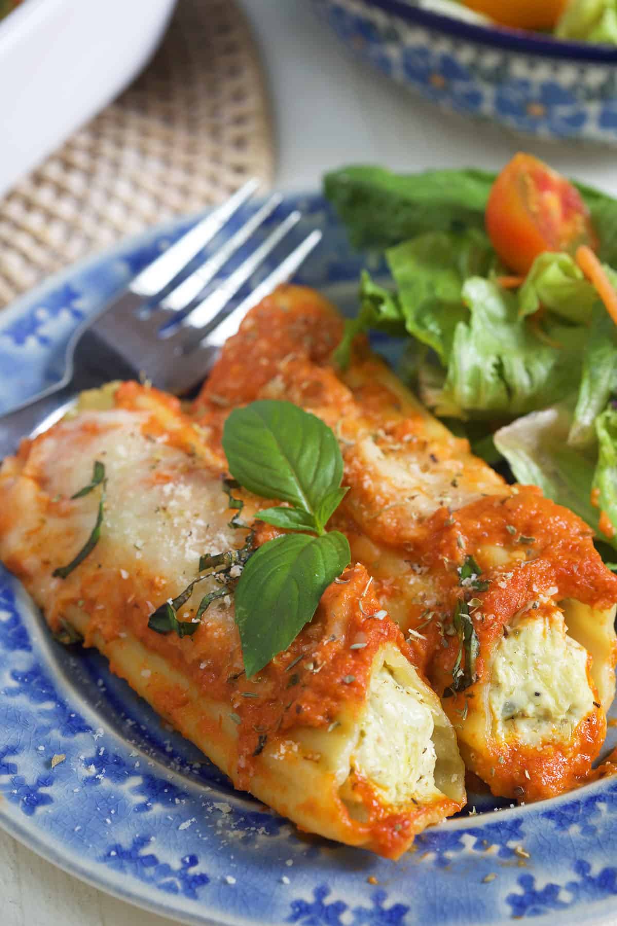 Two pieces of manicotti are placed on a plate next to a fork and side salad.