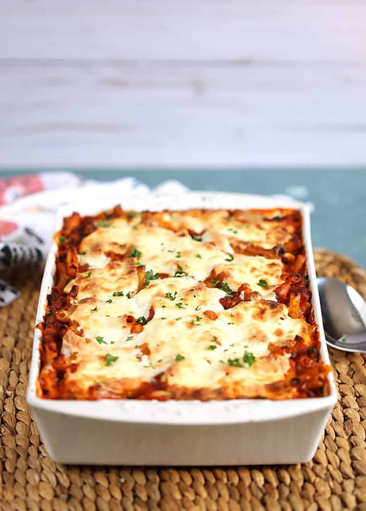 Baked Ziti recipe with melted mozzarella on top in a white rectangular baking dish on a wicker placemat on a blue background 