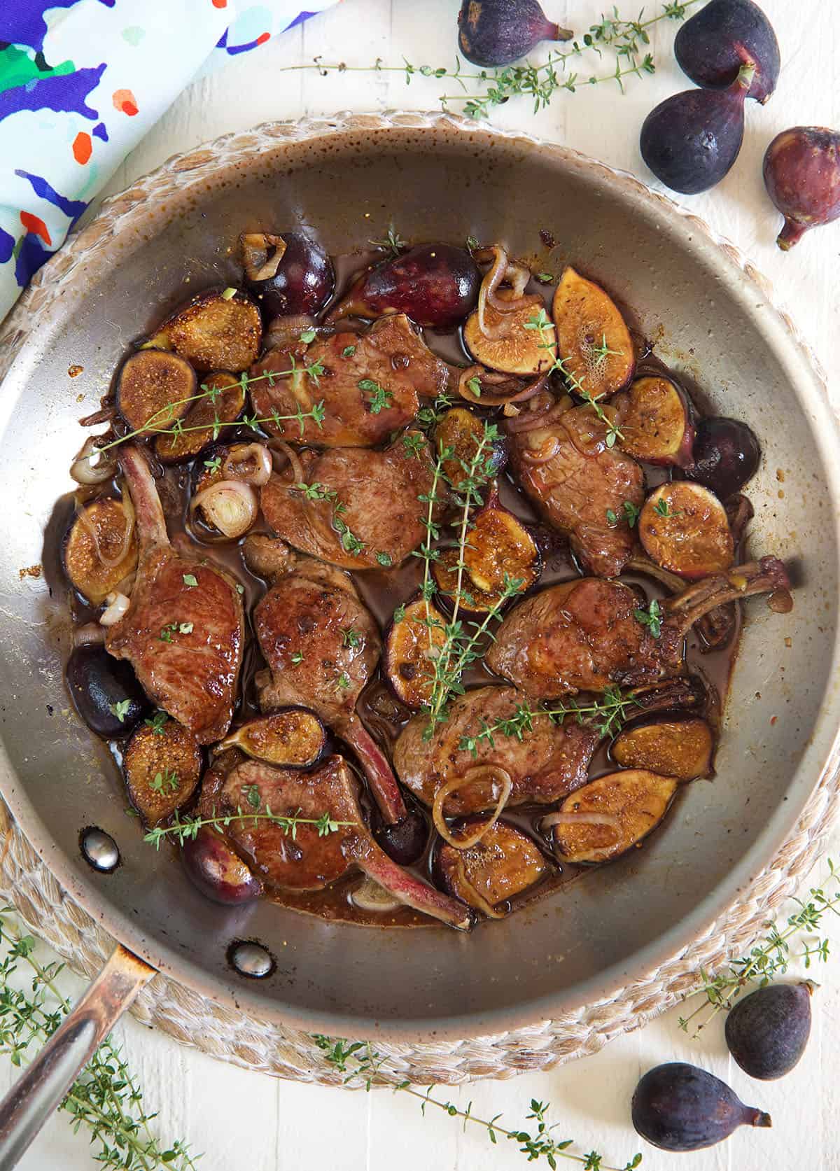 Lamb, figs, and herbs are cooking in a large metal pan. 