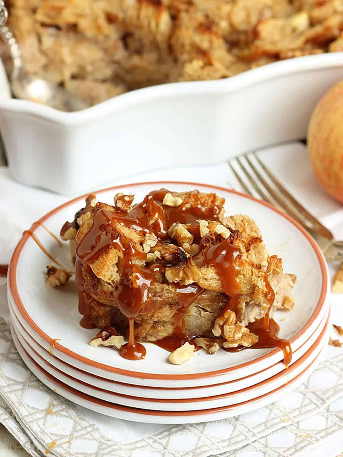 apple bread pudding drizzled with caramel sauce on a stack of white plates.