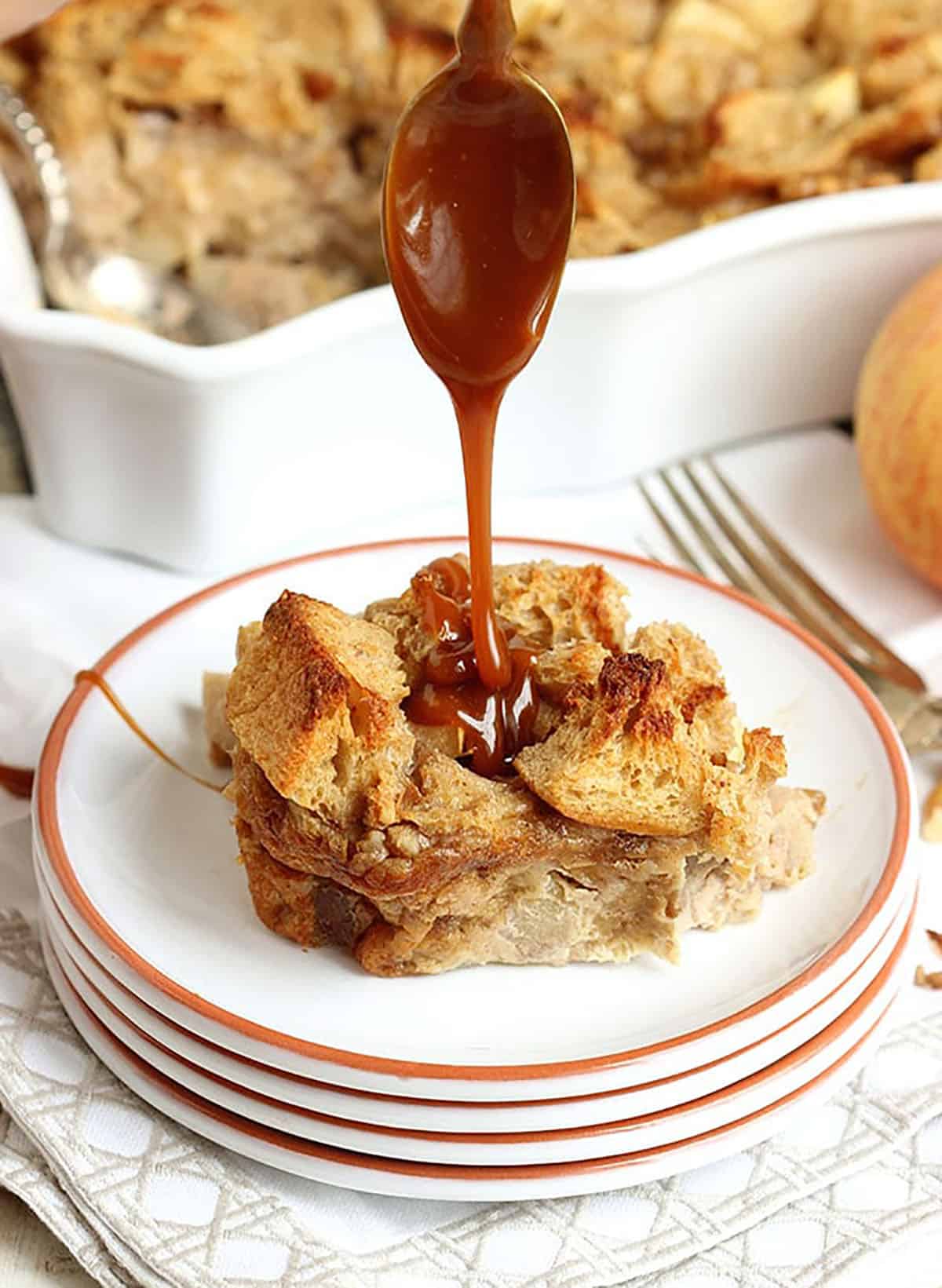 a slice of caramel apple bread pudding being drizzled with caramel sauce with a spoon.