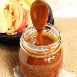 Caramel sauce in a glass jar with a spoon drizzling caramel into it with a plate of apples in the background.