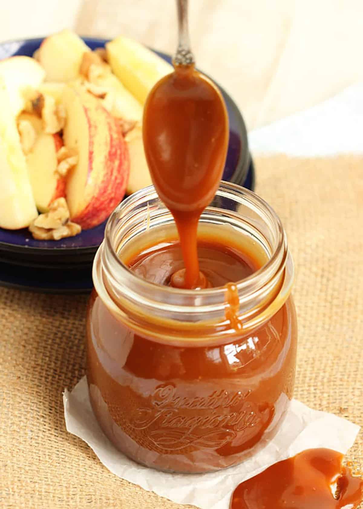Caramel sauce in a glass jar with a spoon drizzling caramel into it with a plate of apples in the background.