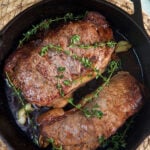 Two steaks are cooked with herbs and butter in a black cast iron skillet.