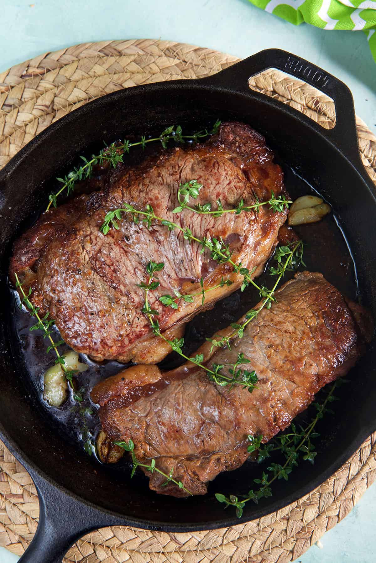 Two steaks are cooked with herbs and butter in a black cast iron skillet.