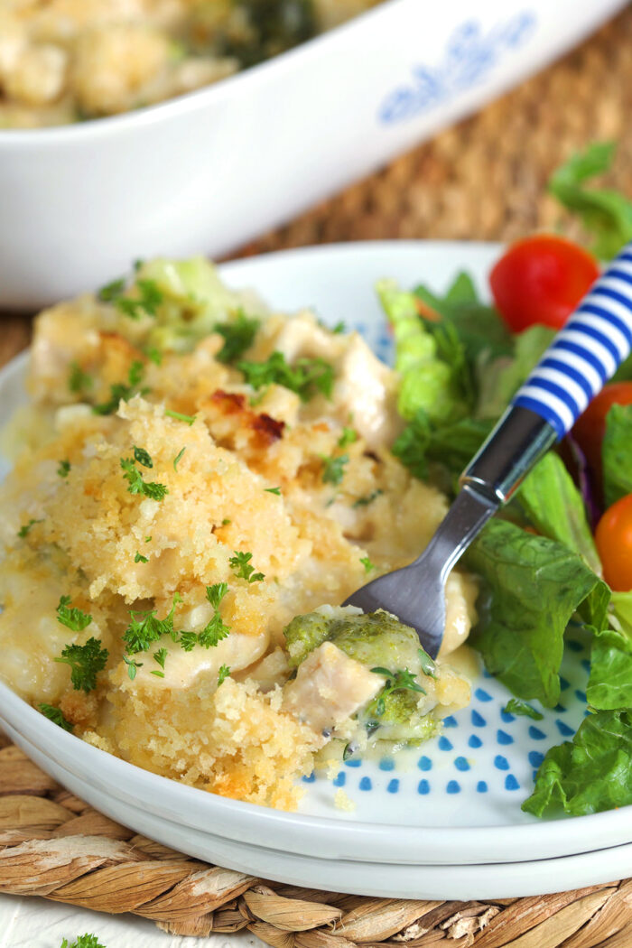 Chicken divan on a white plate with a salad and a blue and white striped fork.