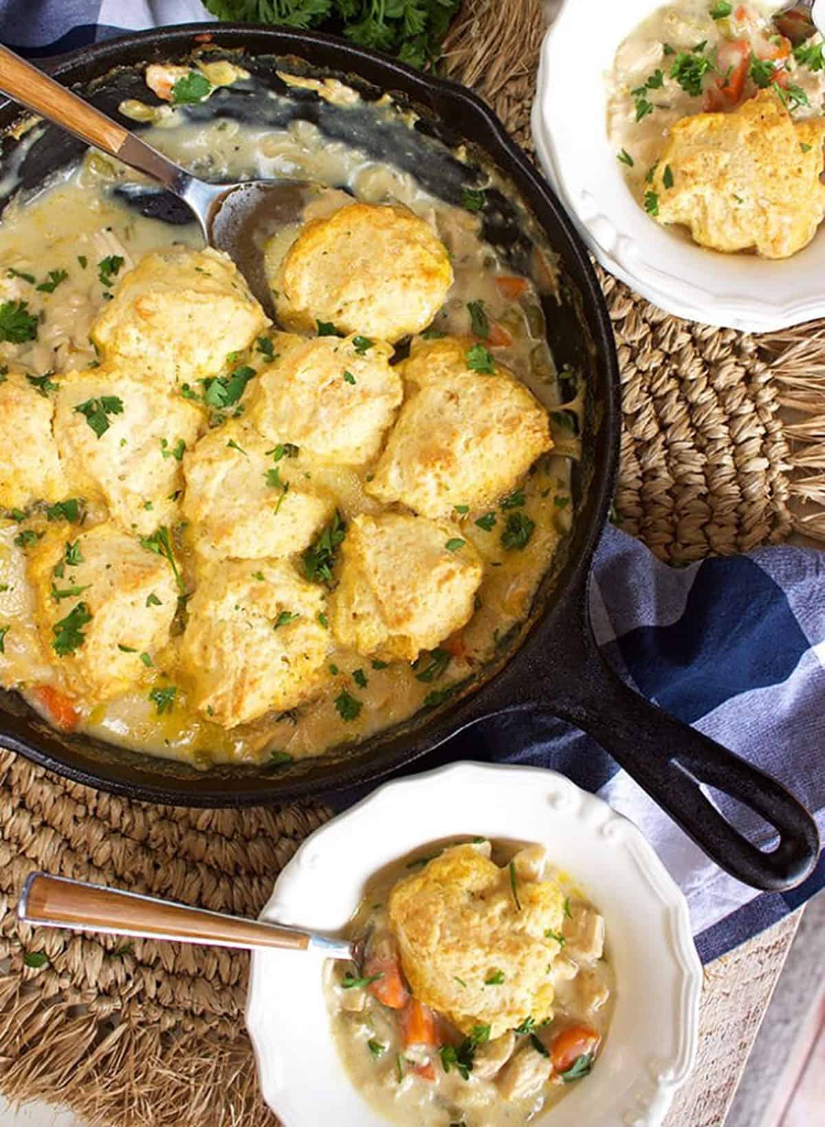Chicken and dumplings in a skillet on a wicker placemat with two white bowls filled with chicken and dumplings.