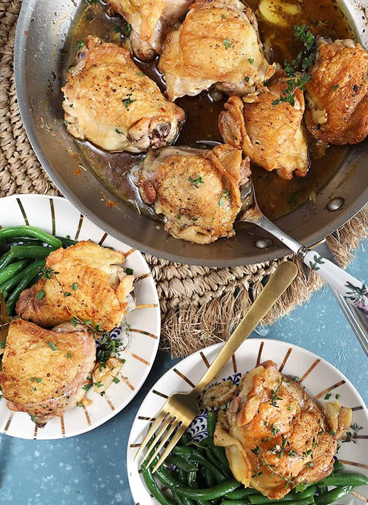 Overhead shot of Crispy Baked Chicken Thighs in a skillet with two white plates with chicken and green beans on a blue background.