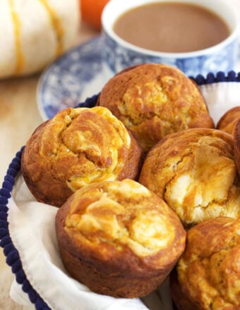 Pumpkin Cream Cheese Swirl Muffins in a basket with a white napkin and a cup of coffee in the background.