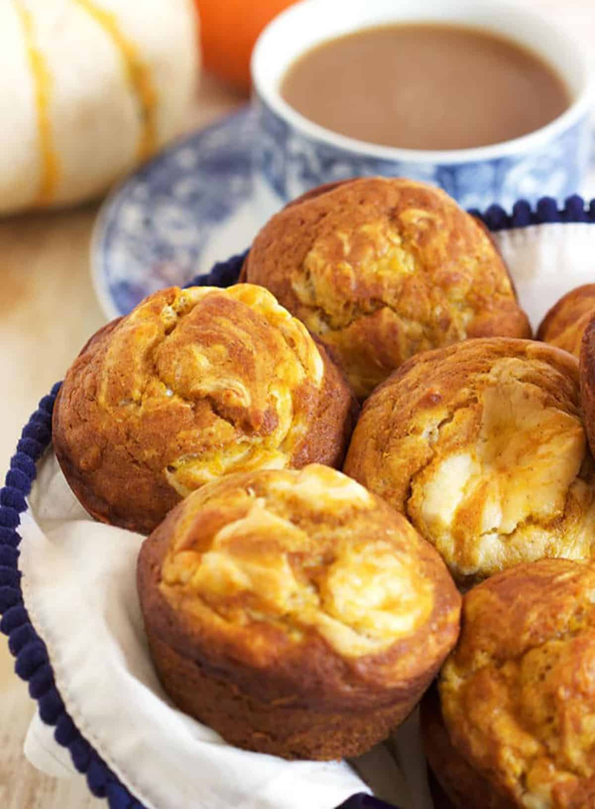 Pumpkin Cream Cheese Swirl Muffins in a basket with a white napkin and a cup of coffee in the background.