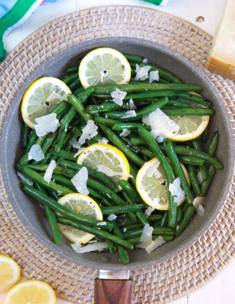 A pan of cooked green beans contains grated parmesan cheese and lemon slices.