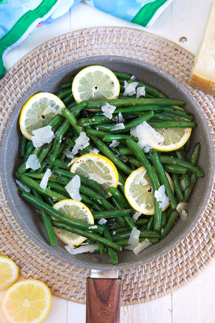 A pan of cooked green beans contains grated parmesan cheese and lemon slices. 