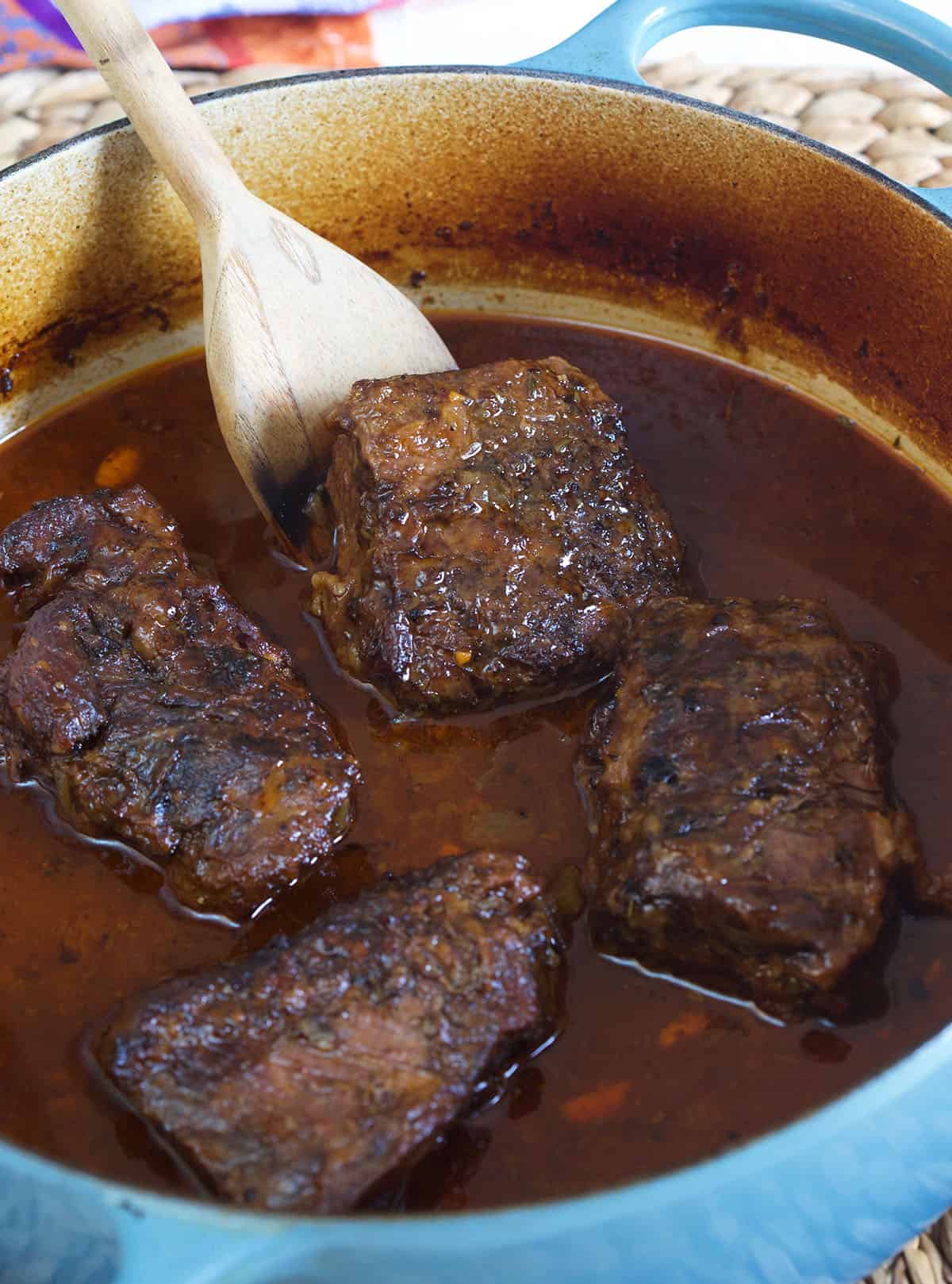A cut of short rib is being moved around with a wooden spoon in a large blue pot.