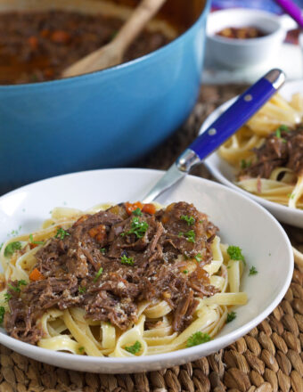 A blue fork is placed in a white bowl filled with noodles and short rib ragu.