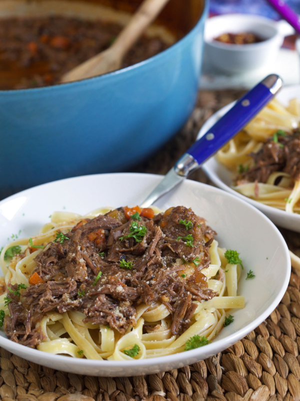 A blue fork is placed in a white bowl filled with noodles and short rib ragu.