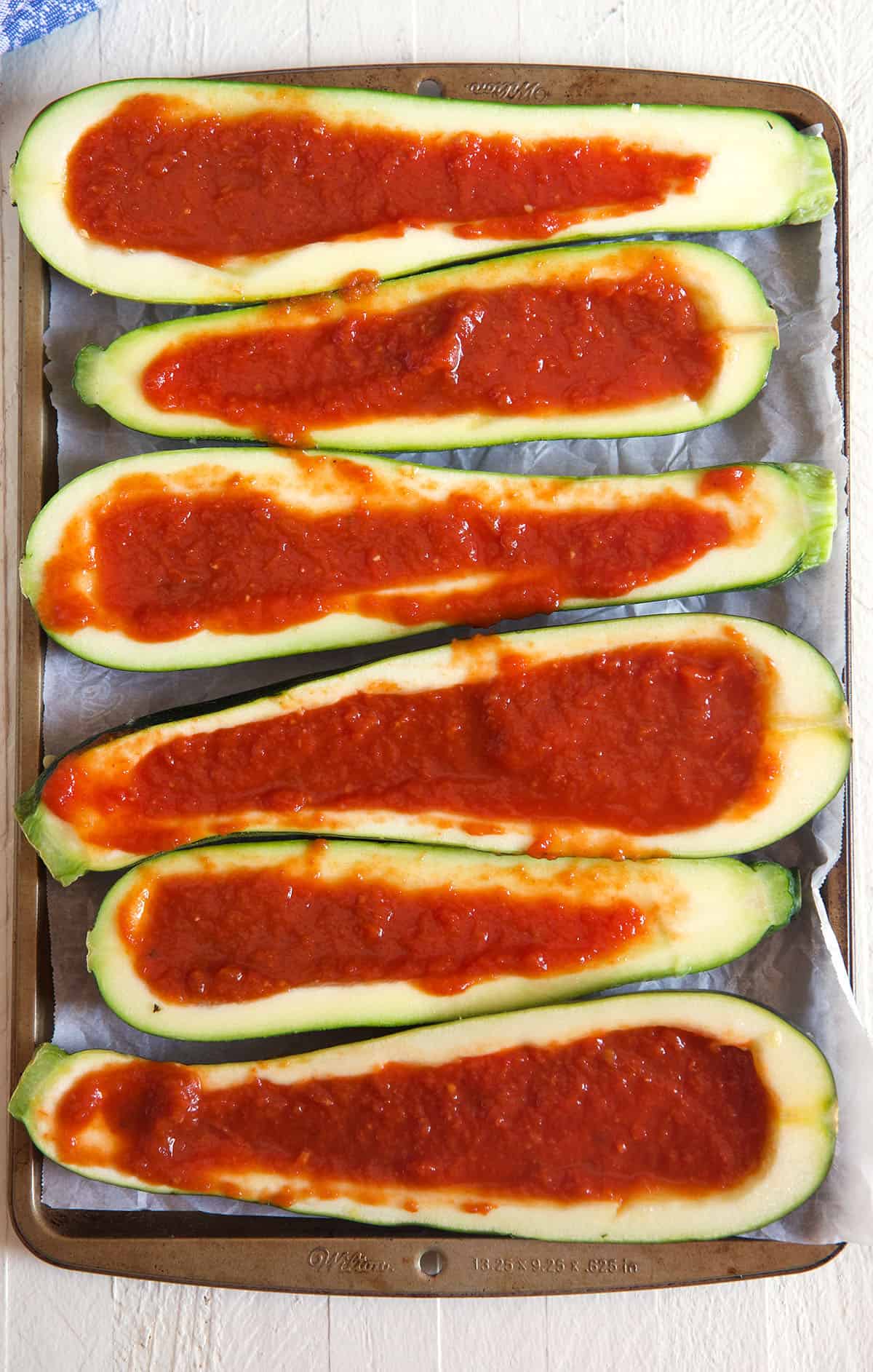 Several sliced zucchini boats are filled with red pizza sauce.