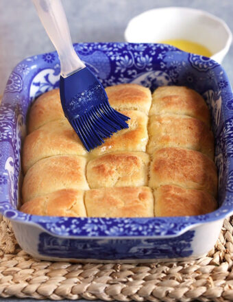 Dinner rolls in a baking dish after baking with a blue brush basting with butter.