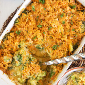 A large silver serving spoon is scooping into a white casserole dish filled with broccoli cheese casserole.