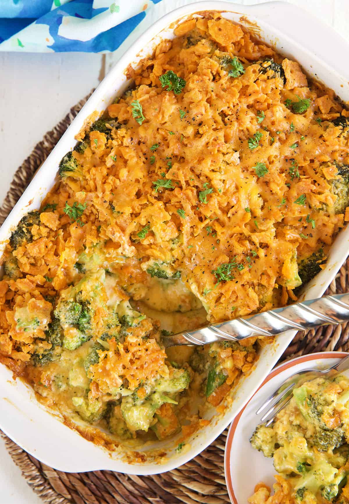 A large silver serving spoon is scooping into a white casserole dish filled with broccoli cheese casserole. 