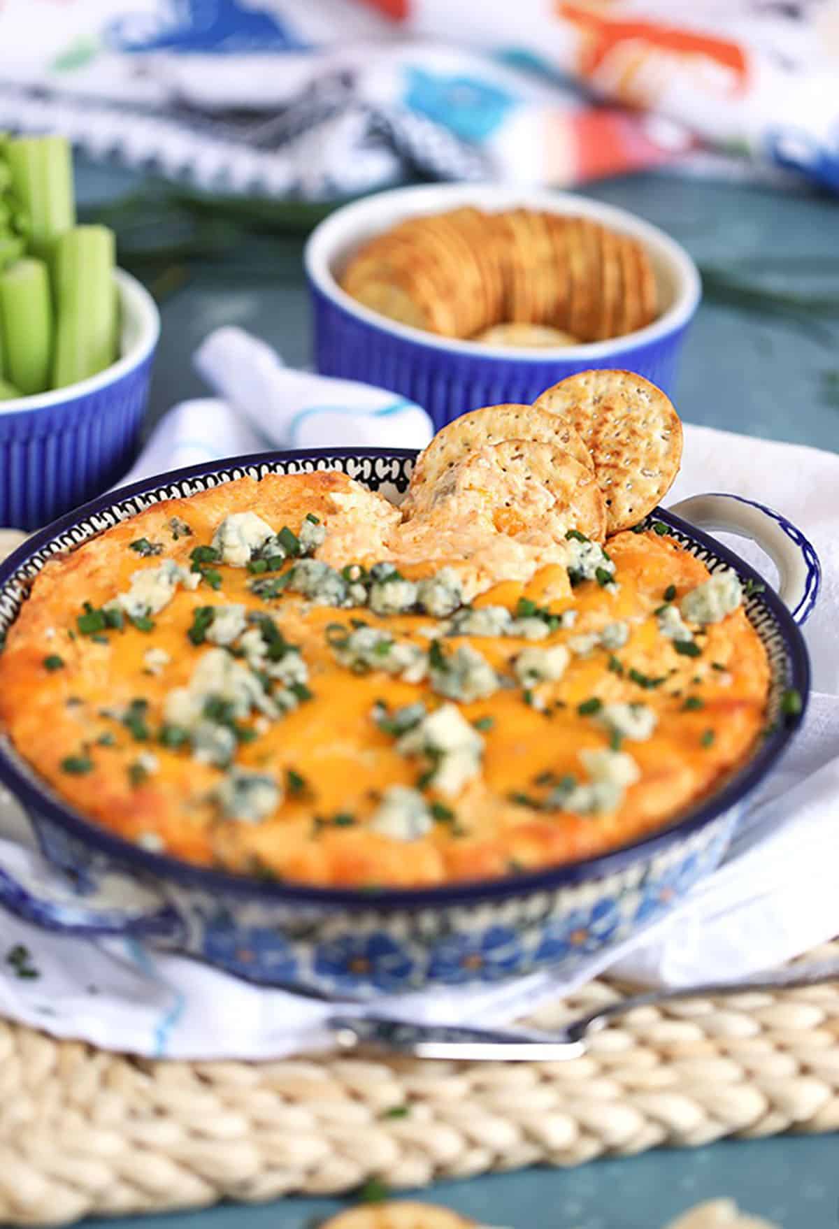 Buffalo Chicken Dip in a floral baking dish on a white napkin on a blue background with crackers and celery. 