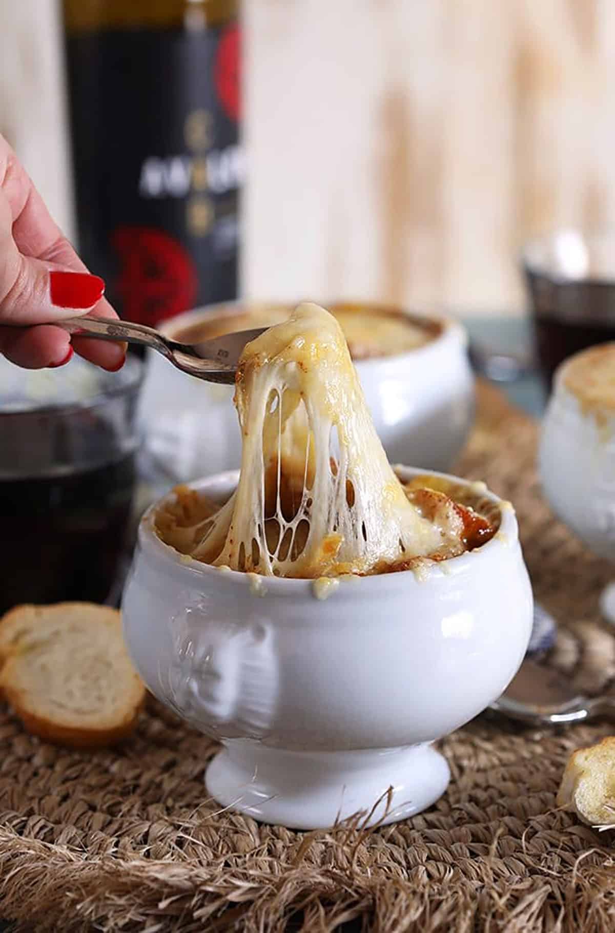 French onion soup in a white crock with a cheese pull on a spoon with a bottle of red wine in the background.