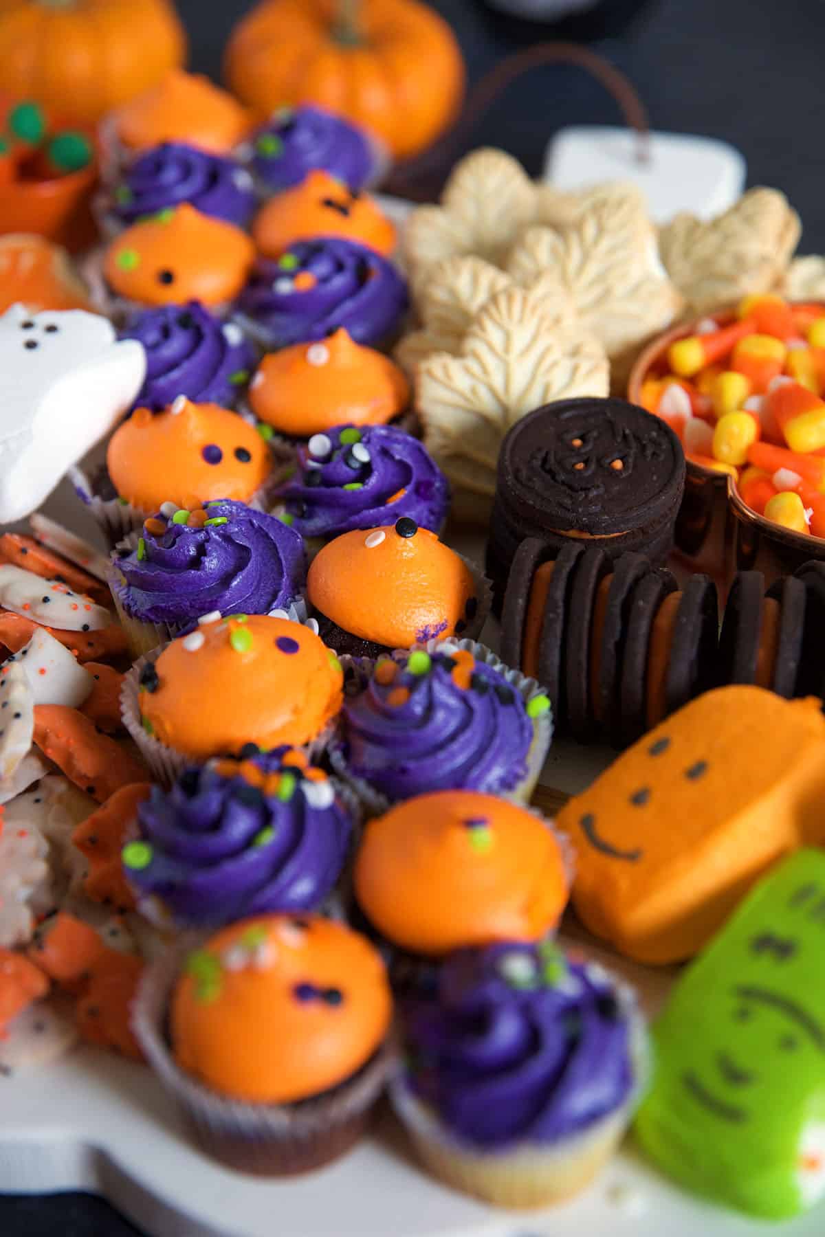 Purple and orange frosted cupcakes are lined up next to other sweet treats on a white pumpkin serving tray.