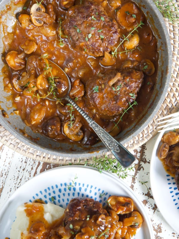 A pan full of hamburger steaks and gravy is placed next to a plate.