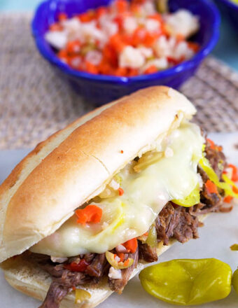 Italian Beef Sandwich on a long roll with a blue bowl of giardiniera in the background.