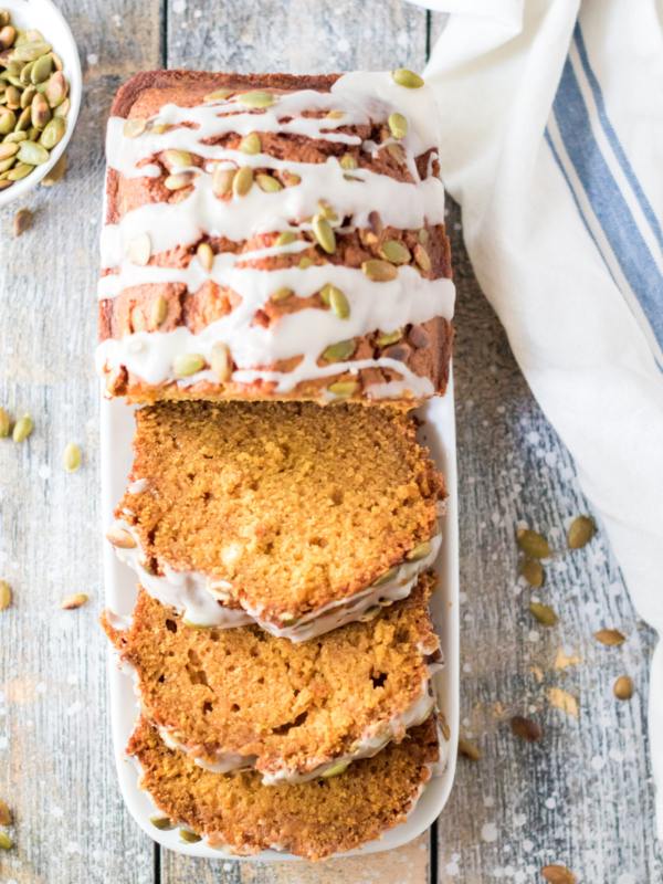 Pumpkin seeds are scattered on a wooden surface around a sliced loaf of pumpkin bread.
