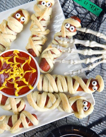 Mummy hot dogs are placed on a white plate with ketchup and mustard in the middle.